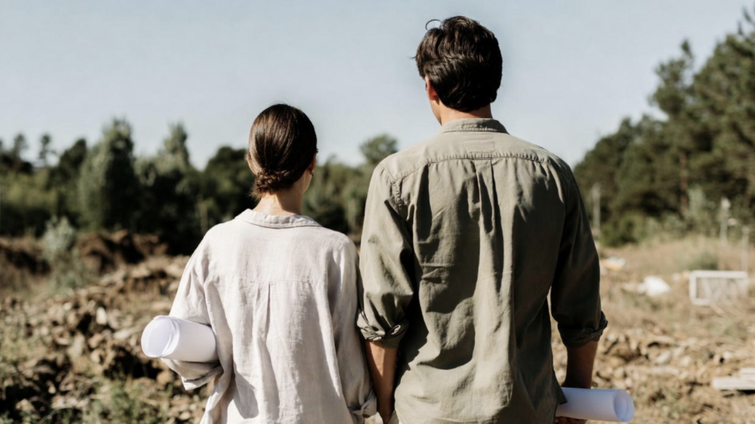 Couple holding house plans while standing on a vacant lot to plan a custom home build